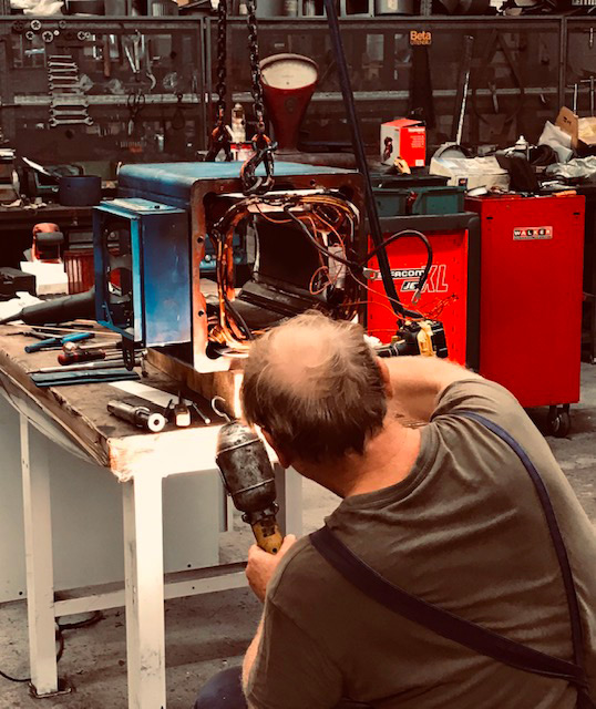 Tecnico con trapano lavora su una carcassa blu di motore elettrico appesa a catene, con fili di rame, in officina
